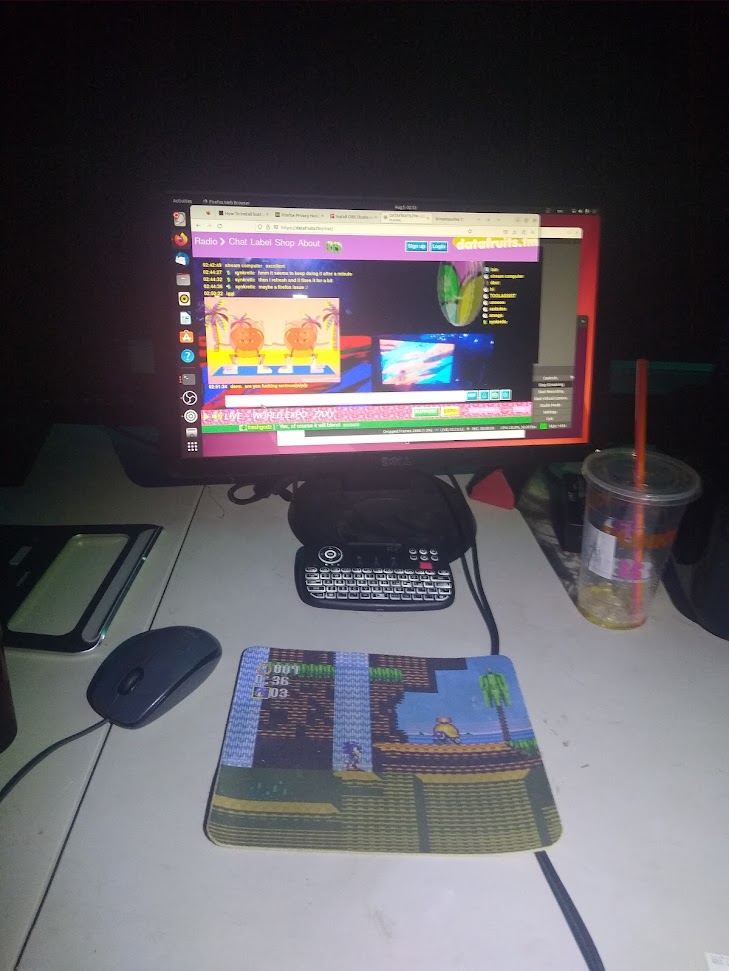 the streaming computer displaying the chatroom in a dark room and my empty iced dunks cup. theres a bluetooth keyboard and a mousepad with a sonic level on it
