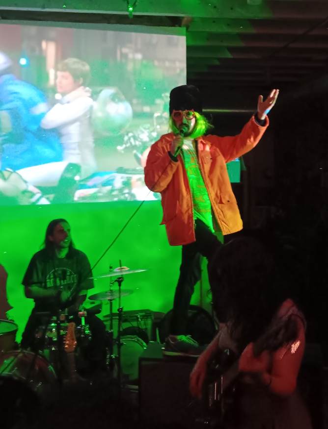Liam in a bright orange jacket, green wig, oversized black beanie, and red large-rimmed glasses, on top of a speaker, singing into a microphone as the fill-in drummer looks on with as much admiration as the crowd